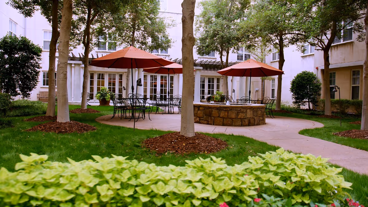 Outdoor courtyard area with green grass, trees, and shrubs. There are several black metal tables and chairs with red umbrellas providing shade. A circular stone fire pit is in the center of the paved area. The courtyard is surrounded by a multi-story building with large windows and white walls.