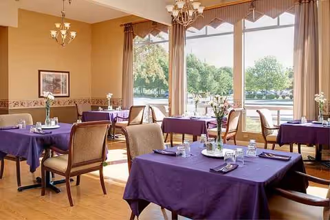 Bright dining room with purple tablecloths and large windows, empty chairs around tables.