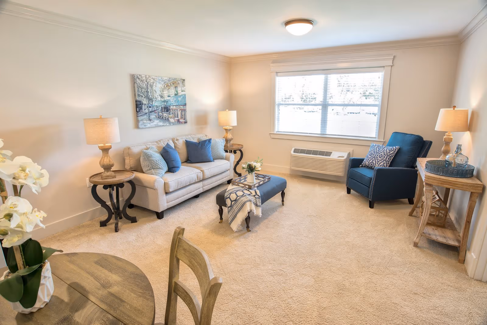 A bright and cozy living room with beige carpet, a beige sofa with blue and beige cushions, a blue armchair with a patterned pillow, two wooden side tables with lamps, a wooden console table with decorative items, a small ottoman with a throw blanket and a tray, a round wooden dining table with chairs, and a large window with blinds letting in natural light.