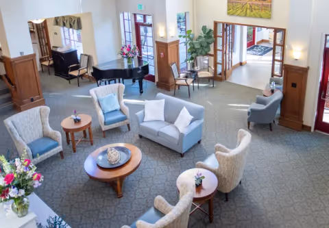 Bright senior living common lounge with sofas, armchairs, coffee tables and a grand piano near the entrance.
