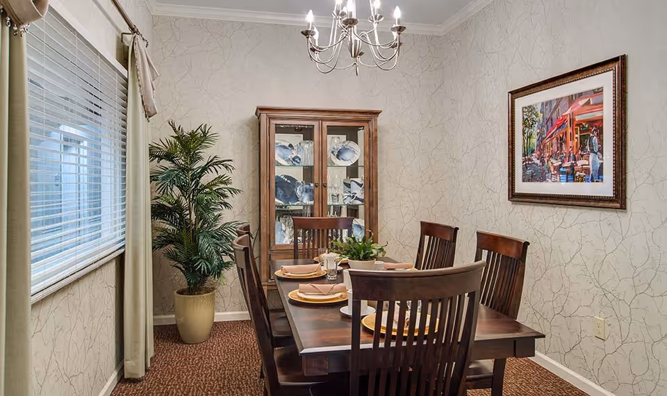 A dining room with a wooden dining table set for six people with plates, napkins, and glasses. There is a wooden china cabinet displaying dishes against the wall, a potted plant in the corner, a window with blinds and curtains on the left, a chandelier hanging from the ceiling, and a framed painting on the right wall.