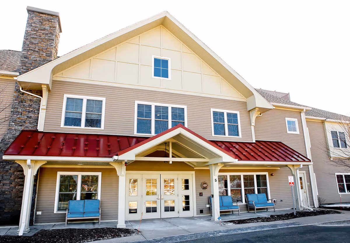 Exterior front view of a senior living facility building with beige siding, a red metal awning above the entrance, multiple windows, and blue benches on the porch area.