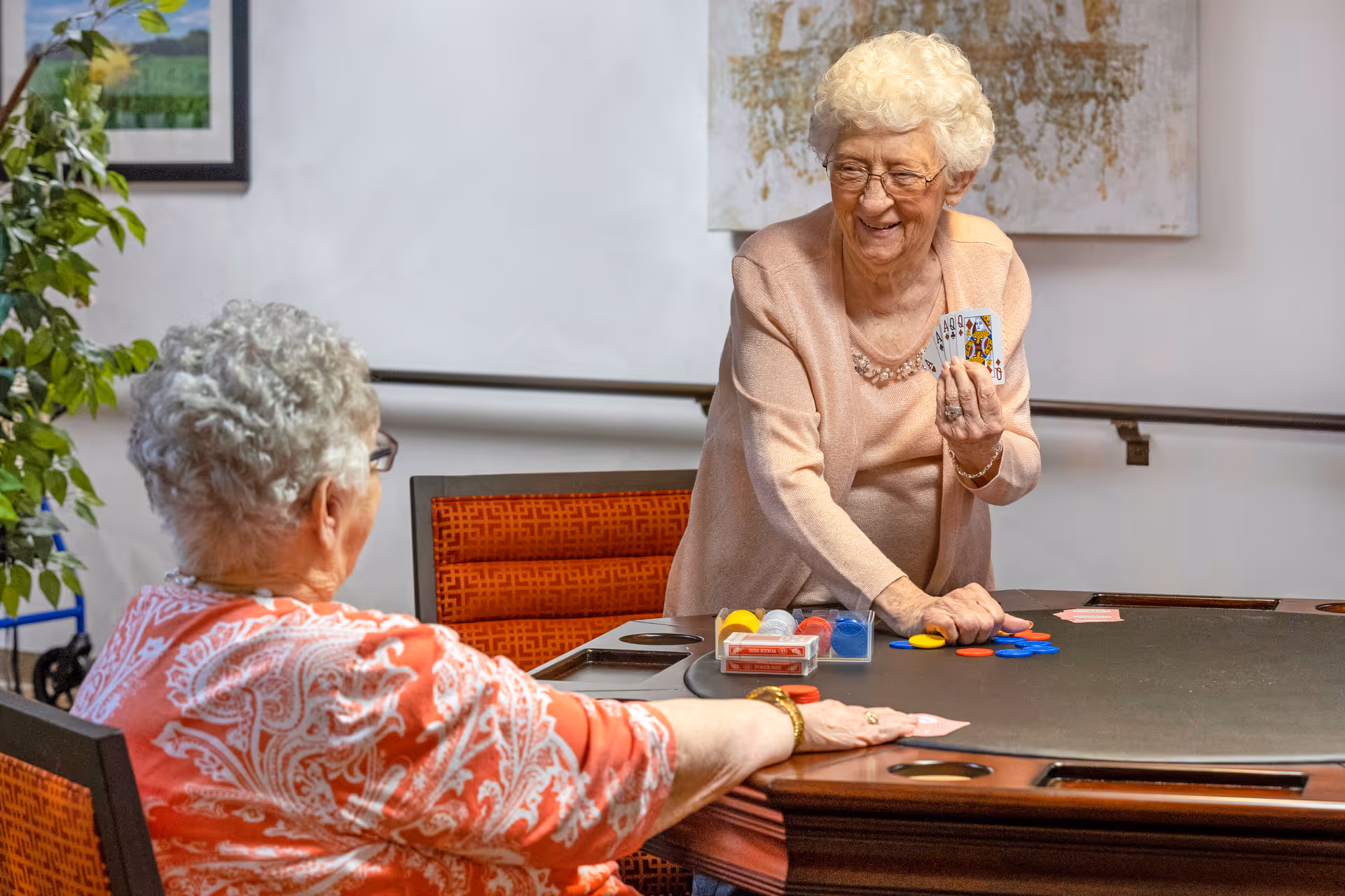 Two elderly women playing cards at a table in a senior living community. One woman is standing and smiling while holding playing cards, and the other woman is seated with her back to the camera. The room has framed artwork on the walls and a potted plant nearby.