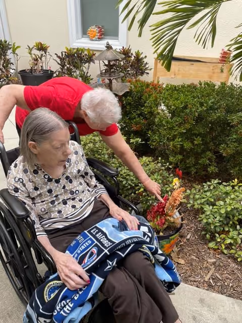 An elderly woman sitting in a wheelchair is covered with a blanket featuring military insignias. Another elderly person in a red shirt is leaning over and pointing at a potted plant with colorful flowers in an outdoor garden area with shrubs and a window in the background.