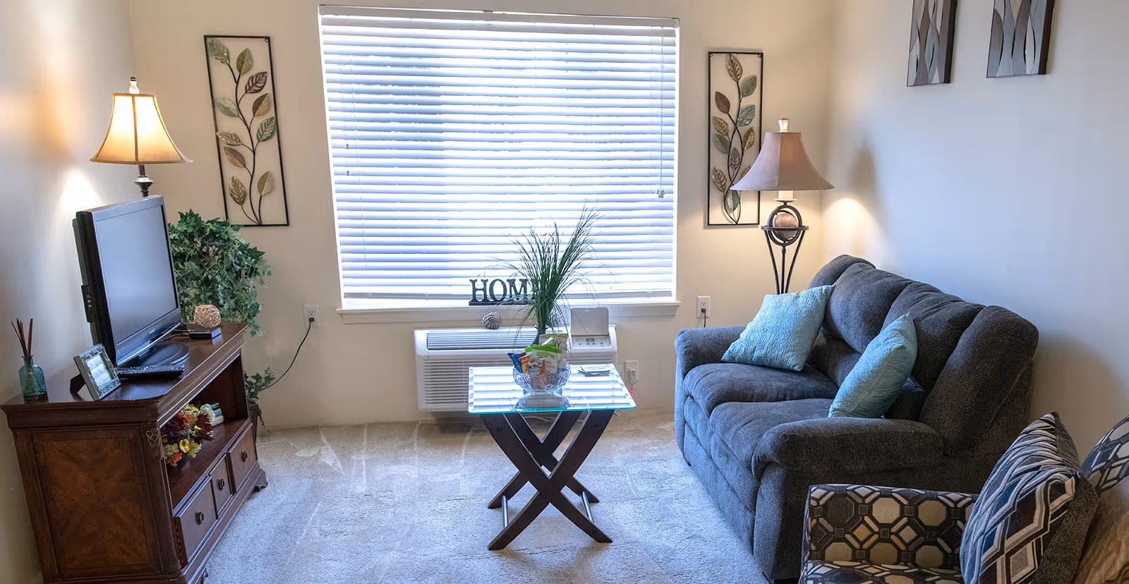 A cozy living room with a gray sofa adorned with blue and patterned cushions, a patterned armchair, a glass coffee table with a plant and snacks, a wooden TV stand with a flat-screen TV, two table lamps, and wall decorations including framed leaf art. A large window with closed blinds lets in natural light.