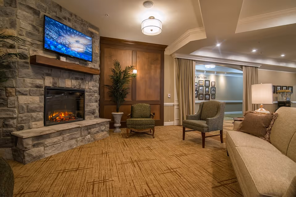 Cozy lounge area with a stone fireplace, wall-mounted TV, upholstered chairs and a sofa under warm lighting.