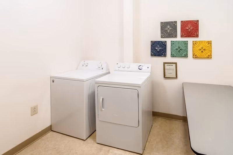 A small laundry room with a white washing machine and dryer side by side against a white wall. Above the machines, there are five decorative square tiles in various colors (gray, red, blue, green, yellow) arranged in a pattern on the wall. A framed notice is also mounted on the wall below the tiles. To the right, there is a gray folding table.