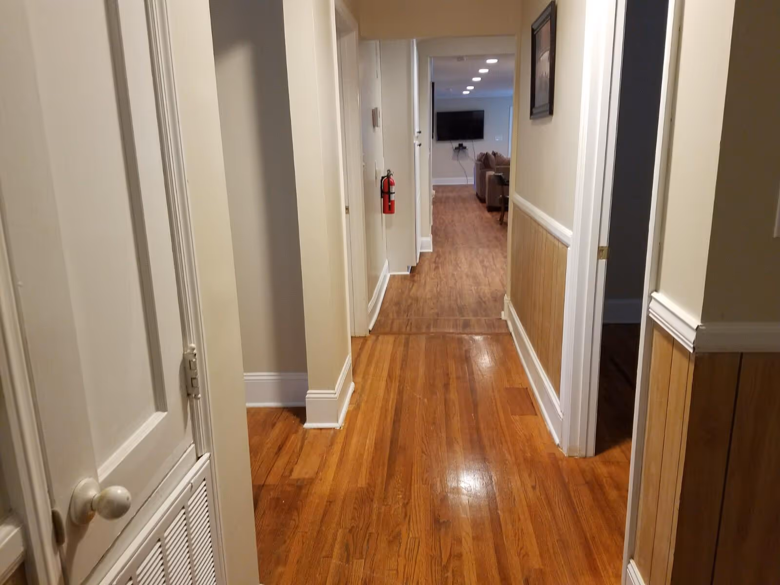 A hallway with wooden flooring and cream-colored walls. There are doors on both sides of the hallway, a fire extinguisher mounted on the left wall, and a living room area with a TV and couch visible at the end of the hallway.
