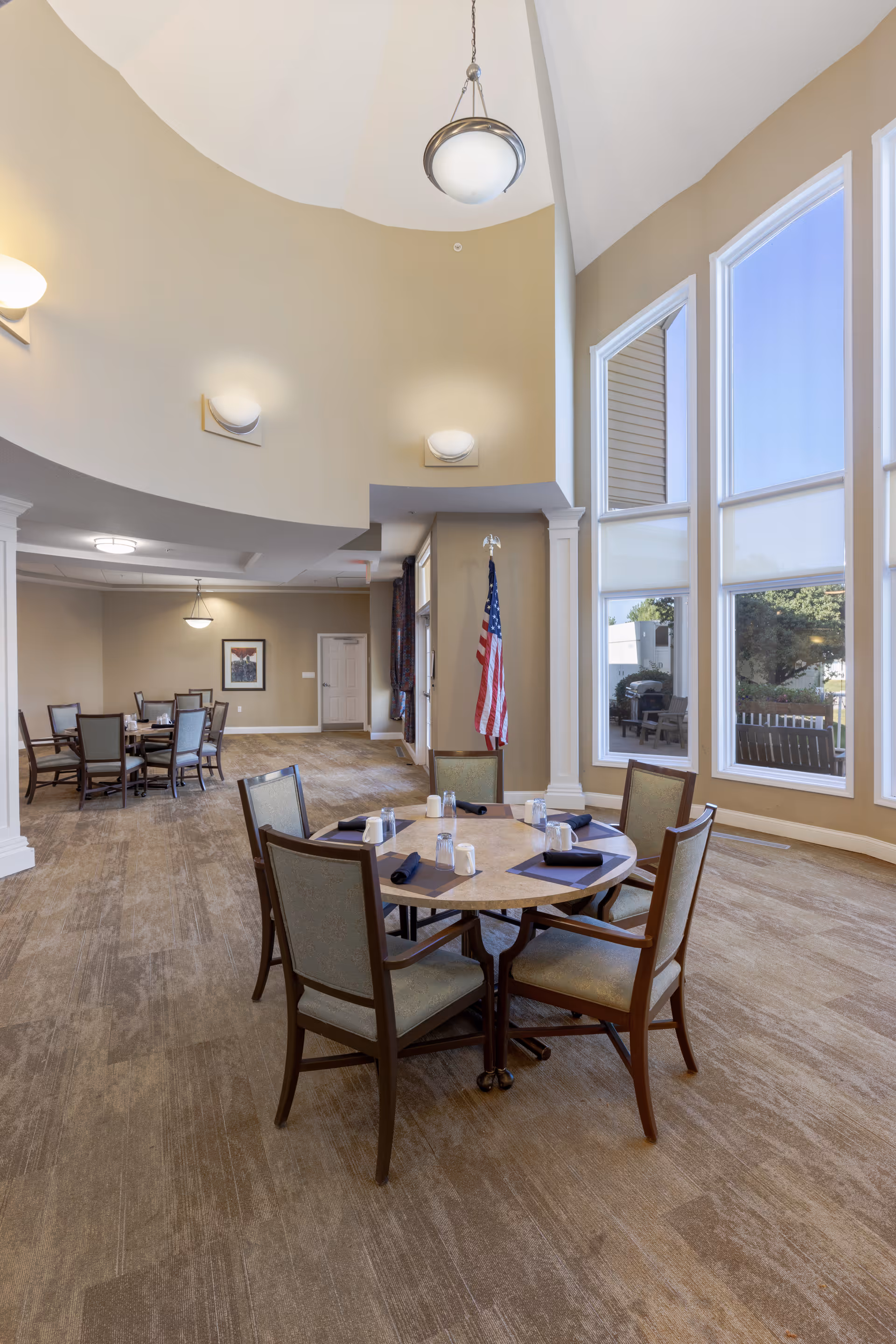 A spacious dining area in a senior living facility with round tables set with napkins, glasses, and salt and pepper shakers. The room features high ceilings, large windows letting in natural light, beige walls, and carpeted floors. An American flag stands near the windows.