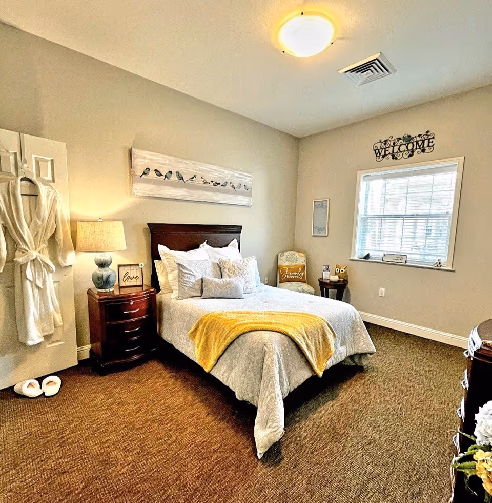A cozy bedroom with a neatly made bed featuring white and gray bedding and a yellow throw blanket. There is a wooden nightstand with a lamp and a decorative sign that says 'love'. A white robe and slippers are hanging on the door. The room has beige walls, a window with blinds, a chair with a cushion that says 'Family forever', and a small round table with decorative items. A 'WELCOME' sign is mounted above the window.
