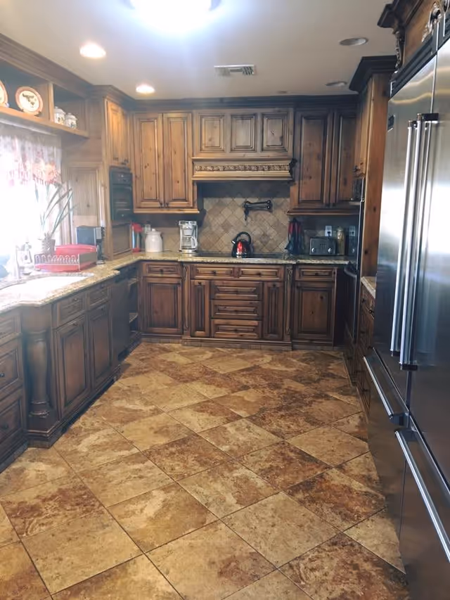 A spacious kitchen with wooden cabinets, granite countertops, and tiled floor. The kitchen features a stainless steel refrigerator, a coffee maker, a toaster, and a kettle on the stove. There is a window with a curtain on the left side letting in natural light.