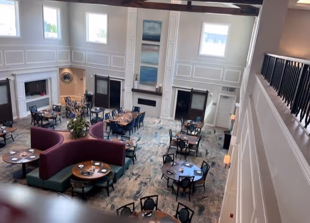 Spacious dining area with multiple round and rectangular tables set with plates and napkins. The room features high ceilings with large windows allowing natural light, white paneled walls, and a patterned carpet. There is a curved purple booth seating arrangement in the center and decorative artwork on the walls.