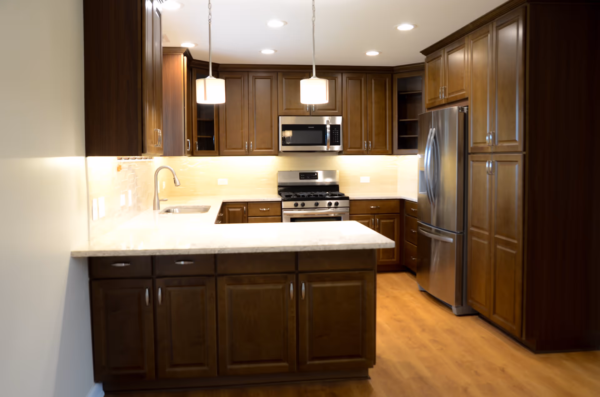 Modern kitchen with dark wooden cabinets, stainless steel refrigerator, microwave, and stove. The kitchen features a white marble countertop island with a sink and two pendant lights hanging above it. The floor is wooden, and the backsplash is light-colored tile.
