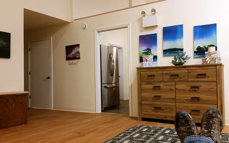 View of an interior living space with wooden flooring, a wooden dresser with decorative items on top, three colorful landscape paintings on the wall above the dresser, and a doorway leading to a kitchen area with a stainless steel refrigerator. A person's feet wearing hiking boots are visible resting on a patterned surface in the foreground.