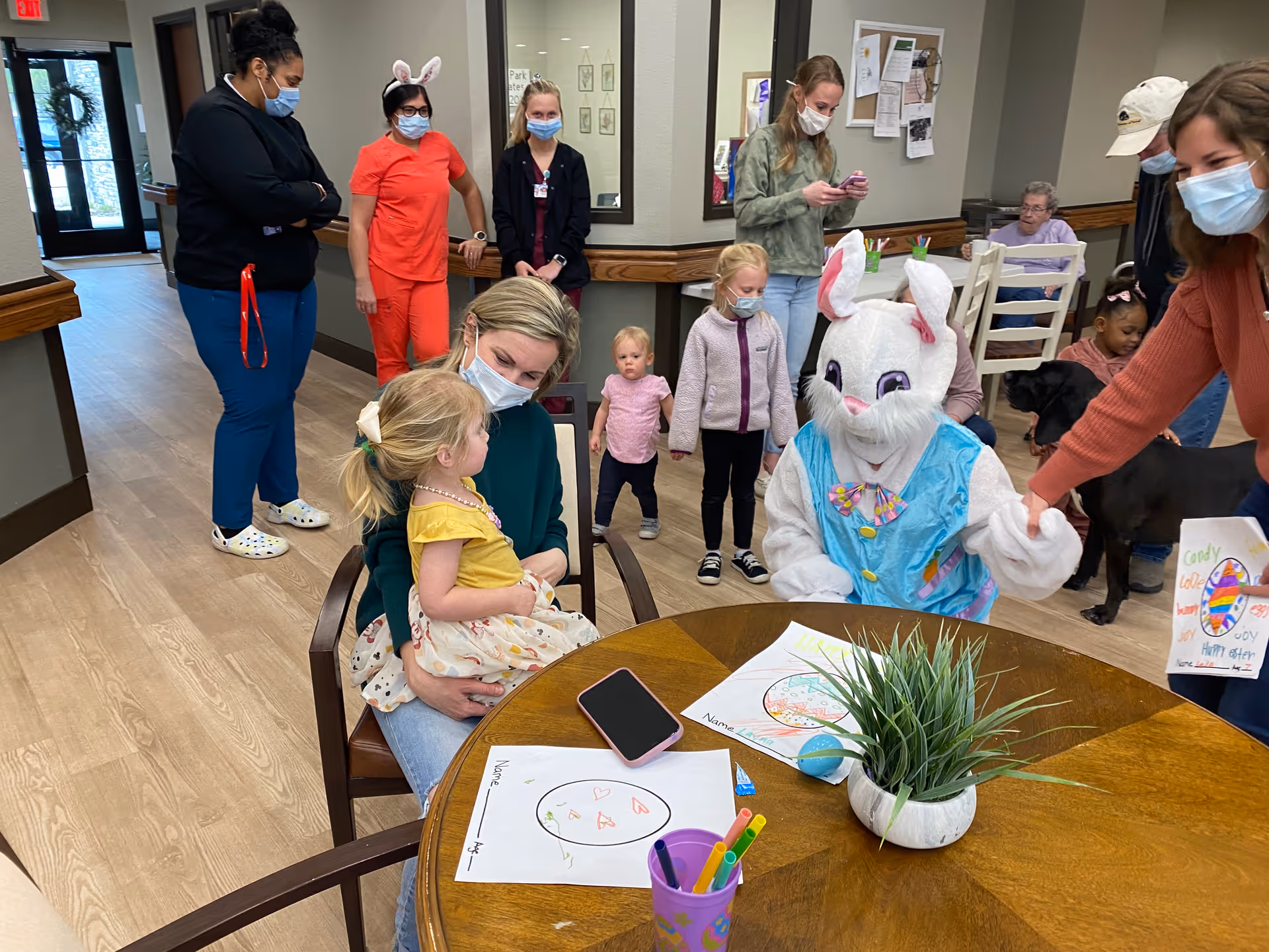 A group of people, including children and adults wearing masks, are gathered in a common area of an assisted living facility. One person is dressed in an Easter Bunny costume sitting near a round wooden table with coloring sheets and markers. A woman is holding a young girl on her lap, and other children and adults are standing or sitting nearby. The setting appears cheerful and festive.