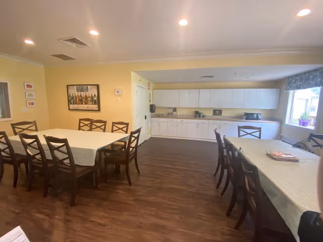 A dining room with two long tables covered with white tablecloths, surrounded by wooden chairs. The room has yellow walls, a wooden floor, and a large window with a floral valance. On one wall, there is a painting and a thermostat. The far wall features white cabinets and countertops with various kitchen appliances like a coffee maker and toaster.