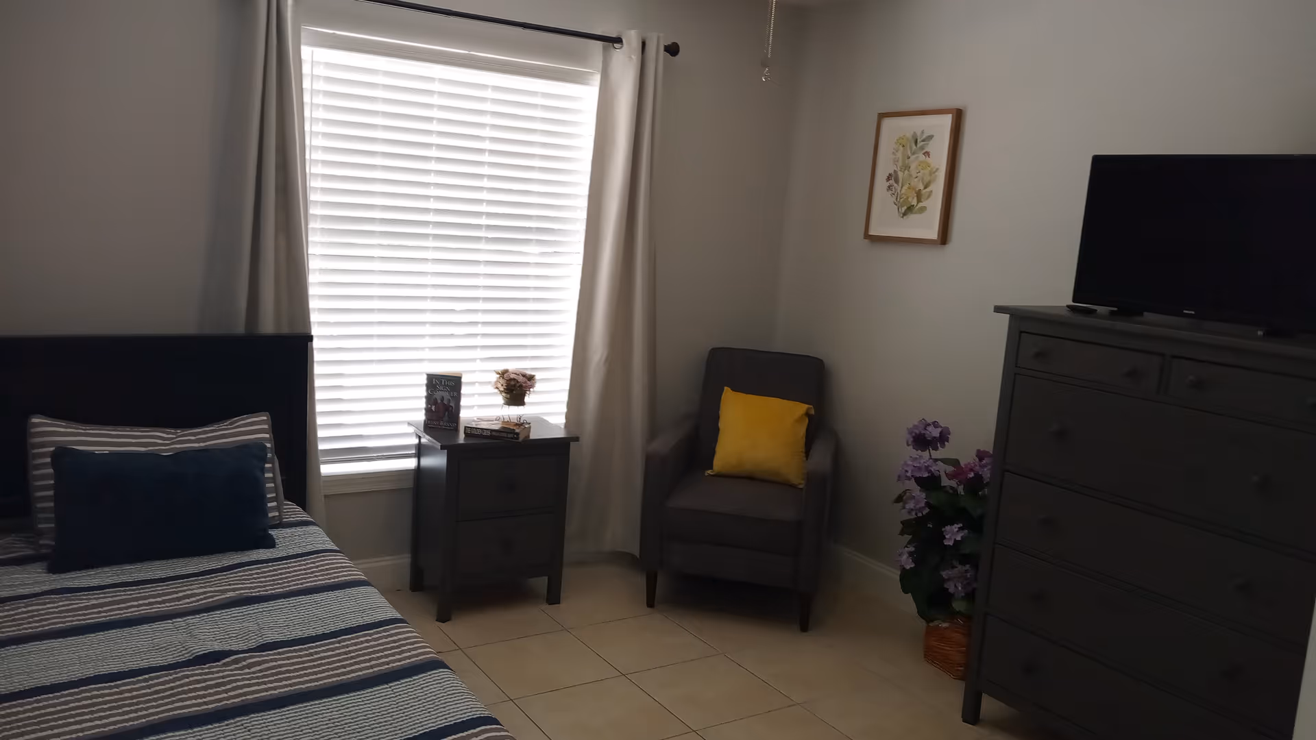 A bedroom with a single bed featuring striped bedding and pillows, a nightstand with books and a small flower vase, a window with closed blinds and curtains, a gray armchair with a yellow cushion, a framed floral picture on the wall, a tall dresser with a television on top, and a potted plant with purple flowers on the floor.