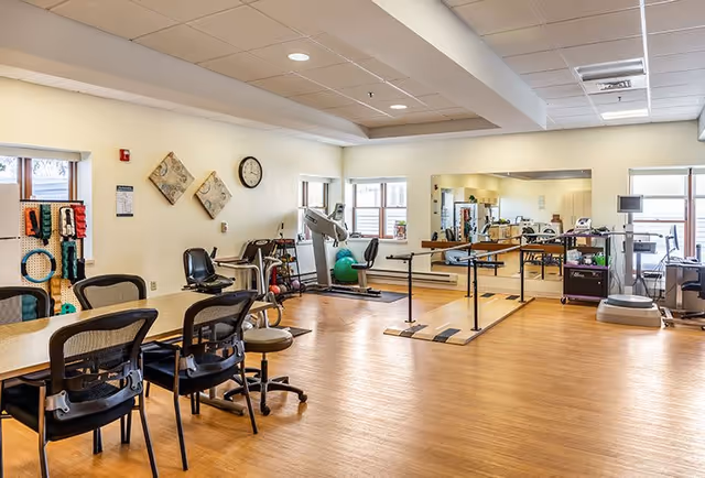 A spacious rehabilitation or physical therapy room with wooden flooring, several chairs around a table, exercise equipment including a treadmill, parallel bars for walking practice, therapy balls, and a large wall mirror. The room is well-lit with natural light coming through multiple windows.