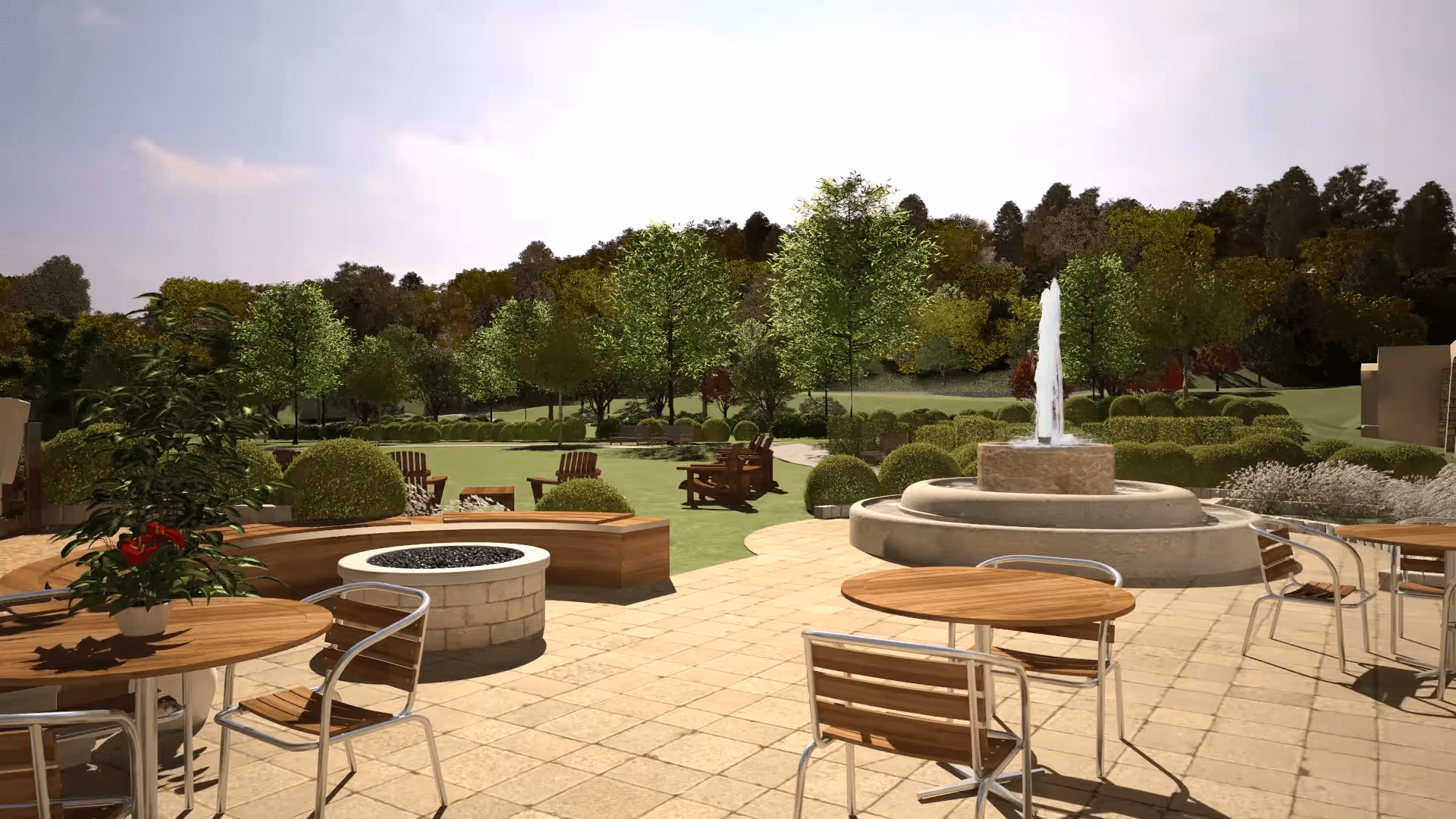 Outdoor patio area with round wooden tables and metal chairs, a circular stone fire pit, a stone fountain with water spouting upwards, surrounded by green trees and bushes under a clear sky.
