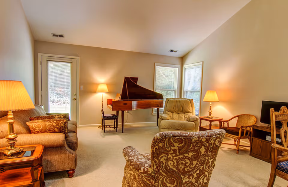 A cozy living room with beige walls and carpet, featuring a brown patterned armchair in the foreground, a beige recliner, a wooden grand piano, a sofa with patterned cushions, two wooden side tables with lamps, a wooden chair, and a small TV on a wooden stand. There are two windows and a glass door letting in natural light.