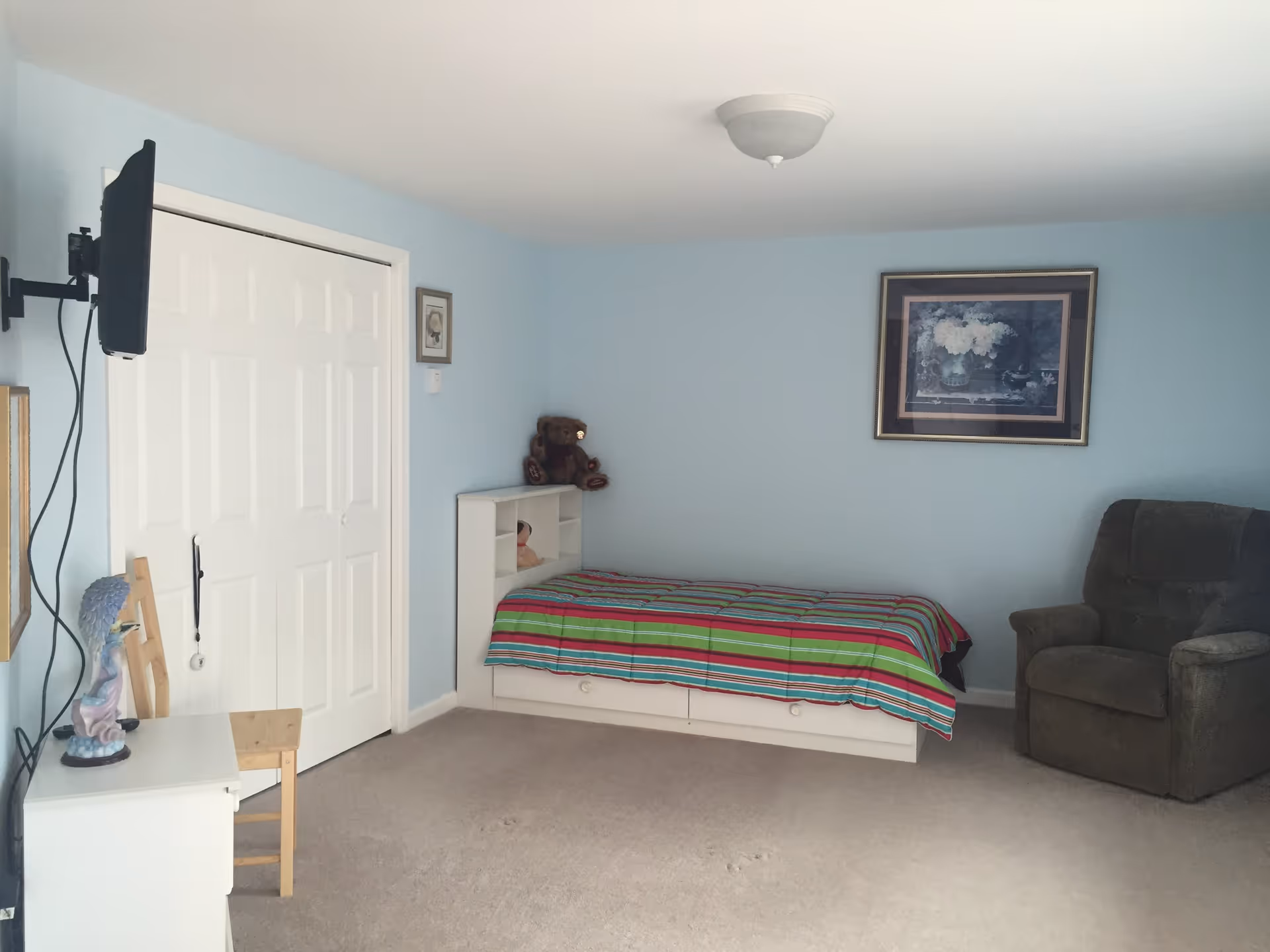 A bedroom with light blue walls featuring a single bed with a colorful striped bedspread, a brown recliner chair, a white dresser with a small statue on top, a wooden chair, a wall-mounted TV, and a closet with white sliding doors. There is a framed picture hanging on the wall above the bed.