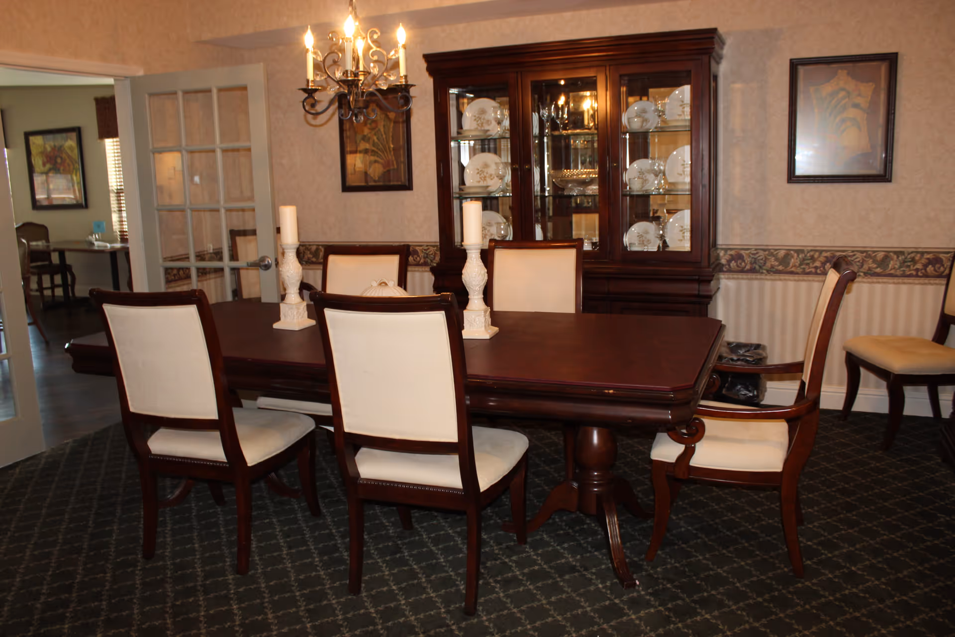 A formal dining room with a dark wooden table surrounded by six cream-upholstered chairs. Two white decorative candle holders with candles are placed on the table. Behind the table is a wooden china cabinet displaying white plates and glassware. The room has patterned wallpaper with a decorative border and framed artwork on the walls. A chandelier hangs above the table, and a glass-paneled door leads to another room.