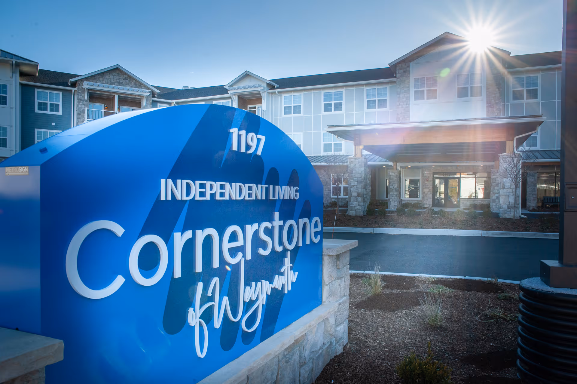 Exterior view of Cornerstone of Weymouth independent living facility with a large blue sign displaying the name and address 1197. The building is a multi-story structure with stone and siding, and the sun is shining brightly in the sky.
