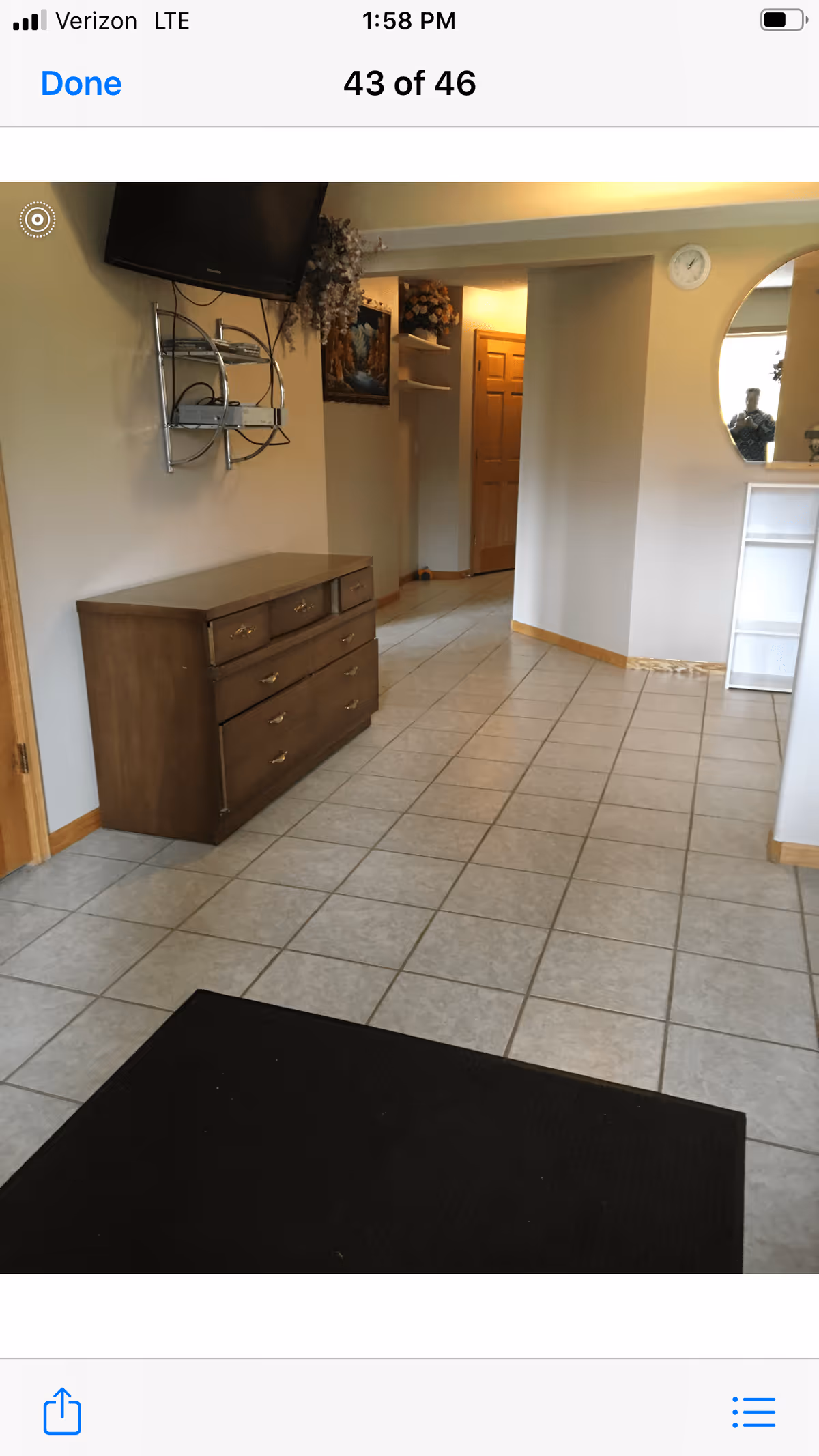 Interior hallway with tiled floor, a wooden dresser with drawers on the left, a wall-mounted TV above the dresser, and a black rug on the floor. The hallway leads to a wooden door and has a small shelf with decorative items on the right wall.