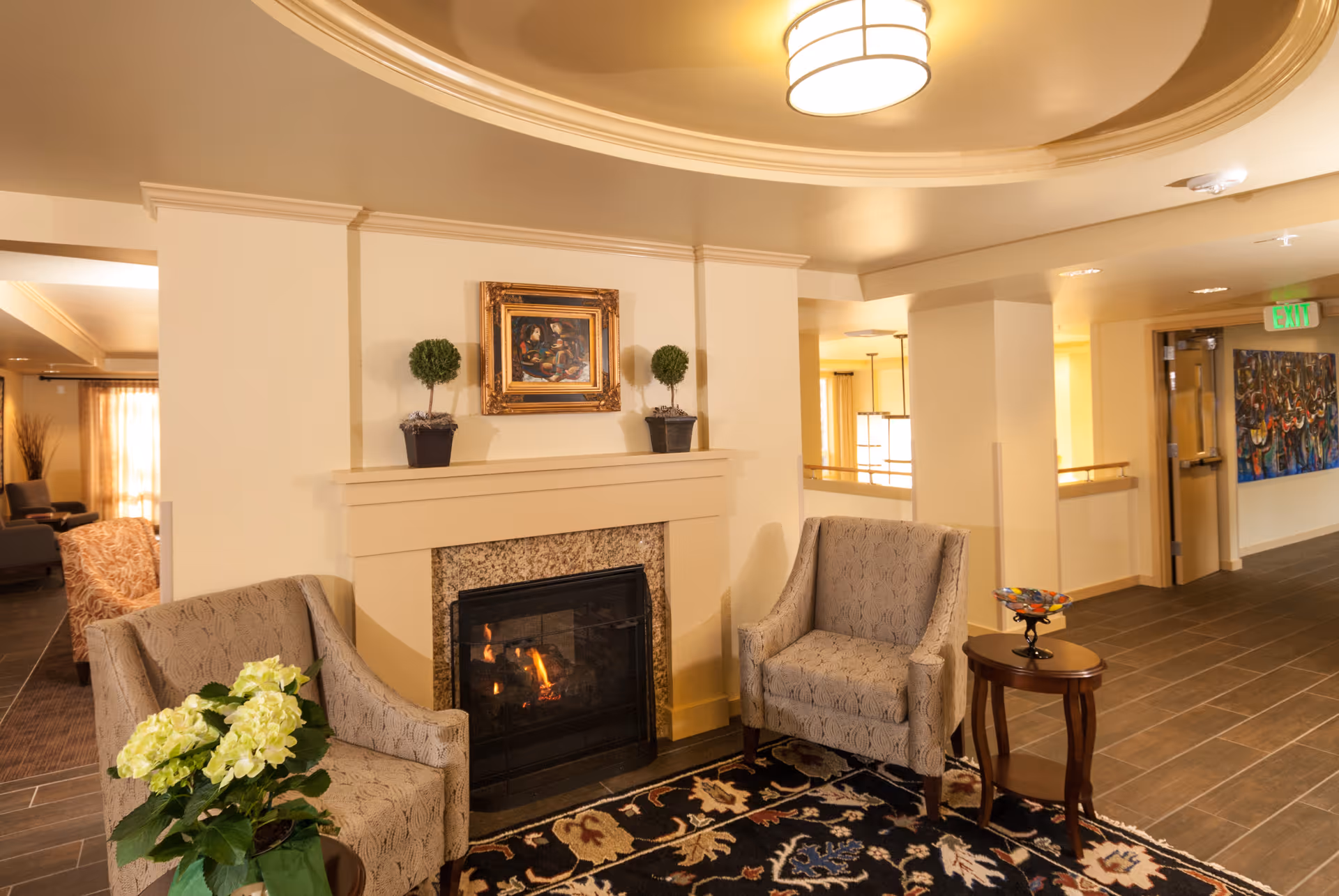A cozy seating area in a senior living facility featuring two patterned armchairs positioned around a fireplace with a fire burning. Above the fireplace is a framed painting and two small potted plants. A small wooden side table with a decorative bowl is placed next to one of the chairs. The area is warmly lit with a ceiling light fixture and has a patterned rug on the floor. In the background, there is a hallway with artwork on the walls and an exit sign.