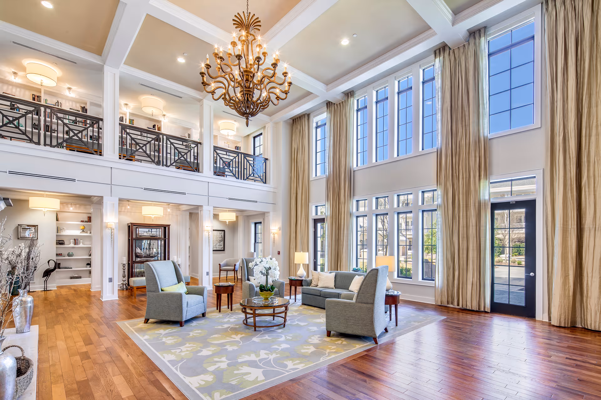 Spacious two-story common room with tall windows, a chandelier, seating arranged on a rug, and an upper balcony.