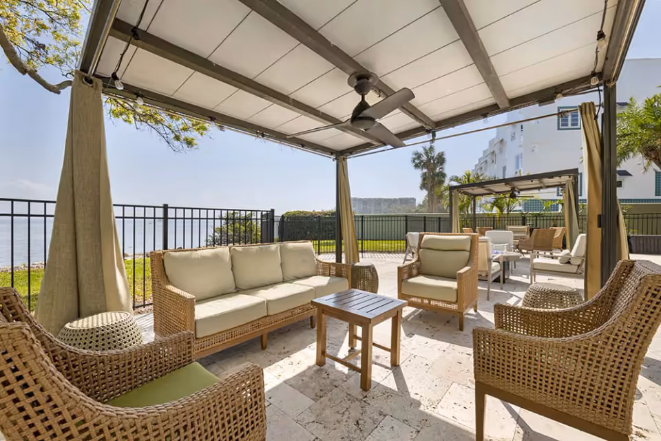 Outdoor seating area with wicker furniture including a sofa, armchairs, and a small wooden table under a canopy with ceiling fans. The area overlooks a body of water and is surrounded by a black metal fence and greenery.