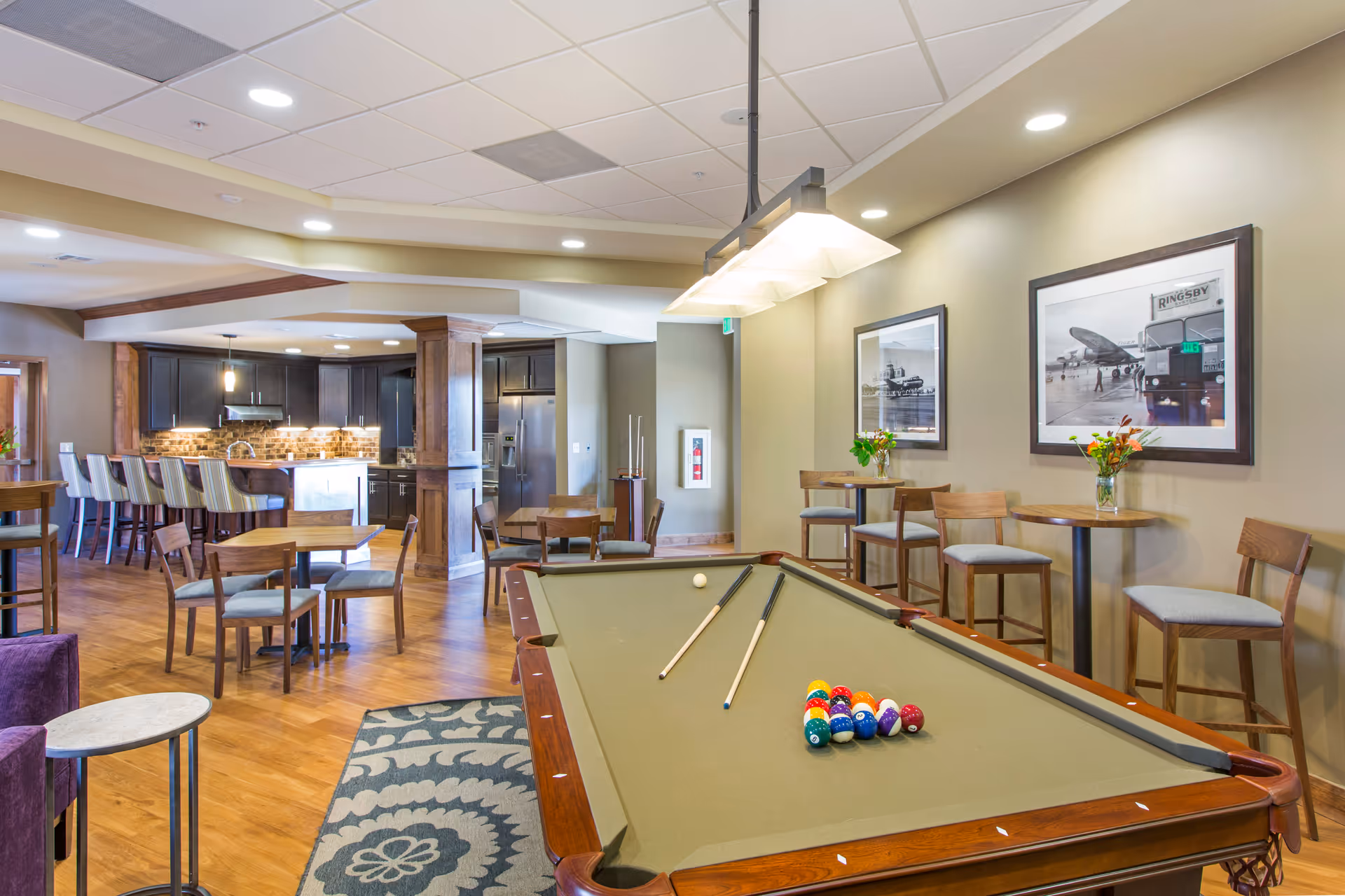 Community lounge with a pool table in the foreground, seating and high-top tables, and a kitchenette/bar area in the background.