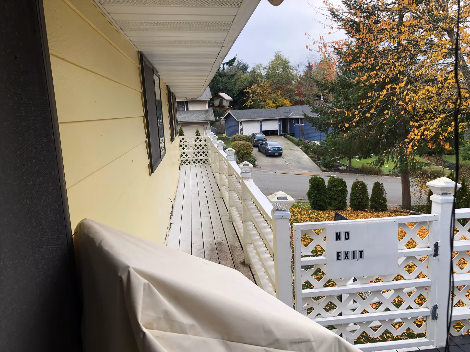 View from a balcony with white wooden railing and a covered object on the left side. The balcony overlooks a street with parked cars, houses, and trees with autumn foliage. A white lattice gate at the end of the balcony has a sign that reads 'NO EXIT'.