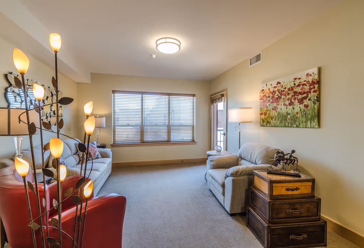 A cozy living room in a senior housing facility featuring a beige sofa, a matching armchair, a red leather chair, a decorative floor lamp with multiple lights, a side table with a lamp, a window with wooden blinds, and a colorful floral painting on the wall.