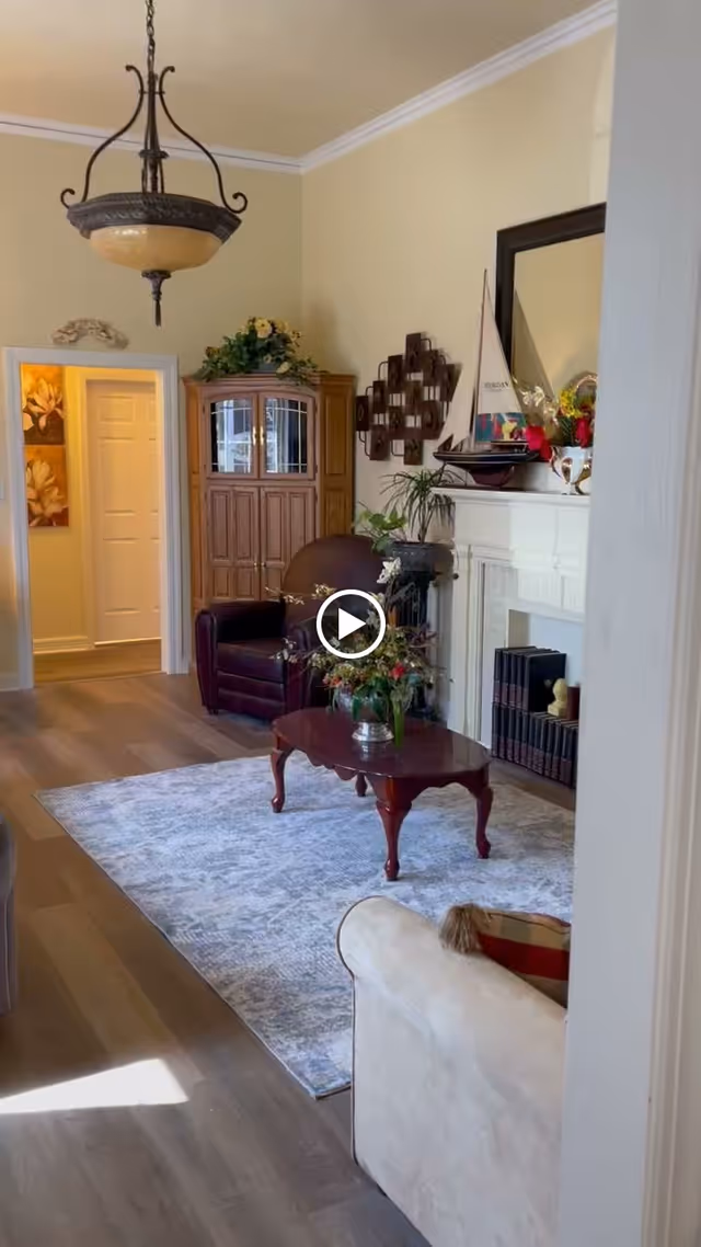 Bright living room with a rug, wooden coffee table, armchairs, fireplace mantel decorated with a model sailboat and plants, and a cabinet.