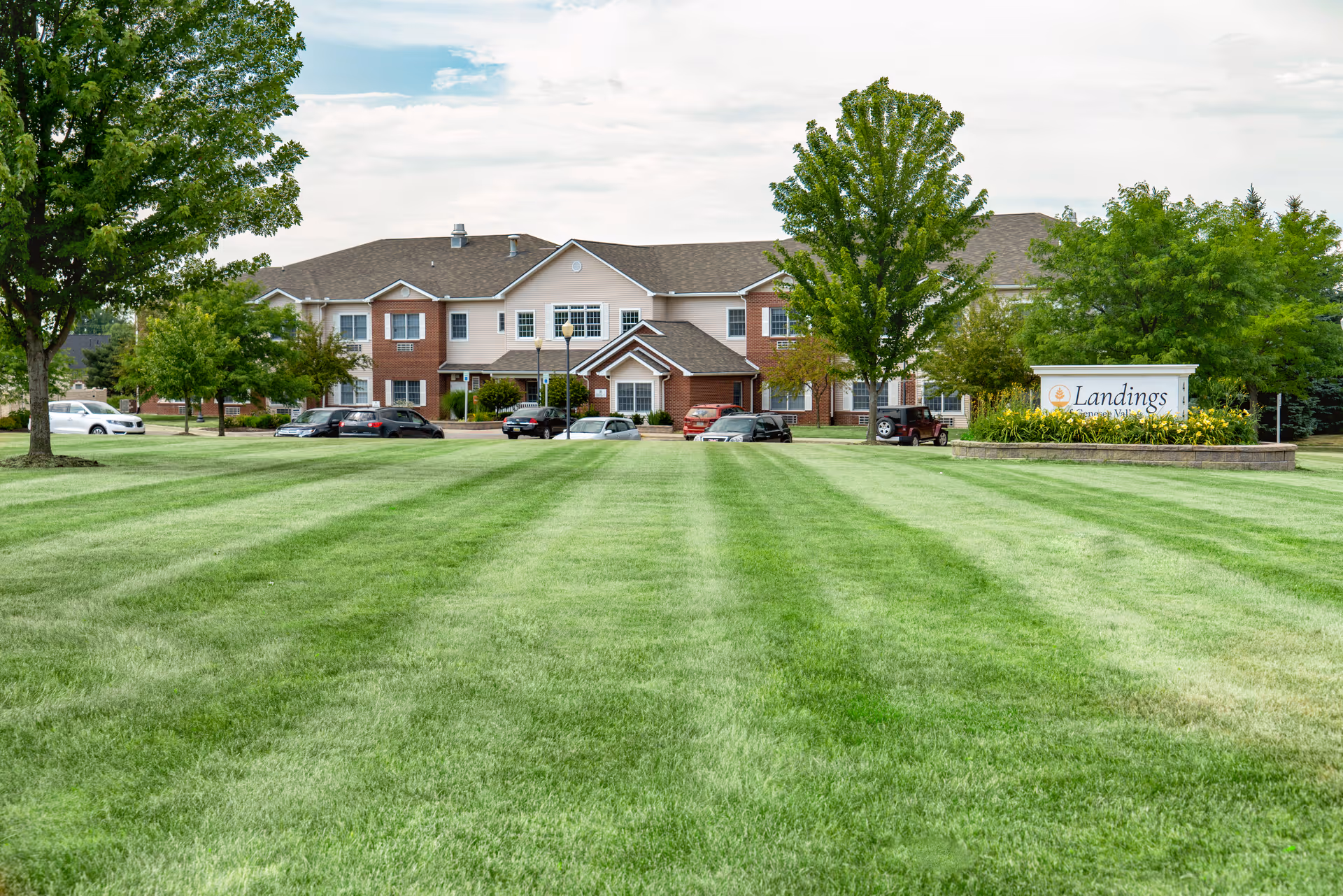 Front view of a two-story senior living building set behind a wide manicured lawn with trees and a 'Landings' sign on the right.