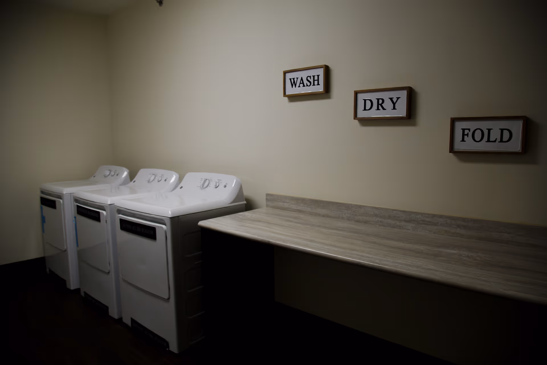A laundry room with three white washing machines or dryers lined up against a beige wall. Above a long countertop on the right side, there are three framed signs on the wall that read 'WASH', 'DRY', and 'FOLD'.