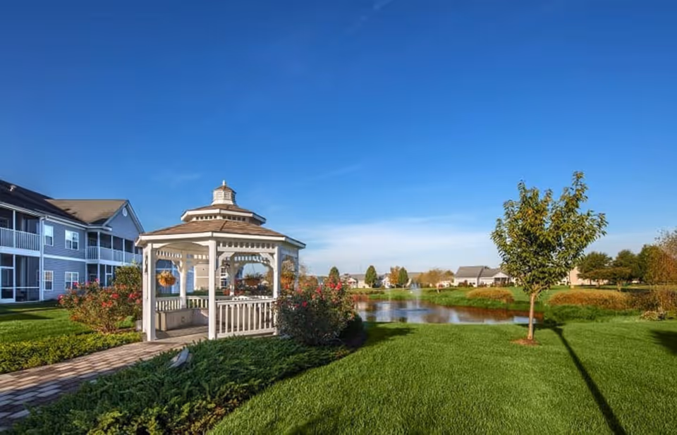 A white gazebo surrounded by green grass, bushes, and a small tree near a pond with a fountain. Residential buildings are visible in the background under a clear blue sky.