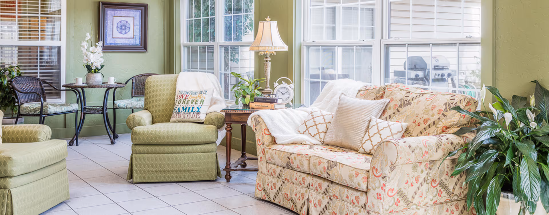 A bright and cozy living room area with a floral patterned sofa adorned with multiple cushions and a white throw blanket. Next to the sofa is a wooden side table with a lamp, clock, and books. There are two green upholstered armchairs, one with a decorative pillow. In the background, there is a small round table with two wicker chairs, a framed picture on the wall, and large windows letting in natural light. Several green plants add a touch of nature to the room.