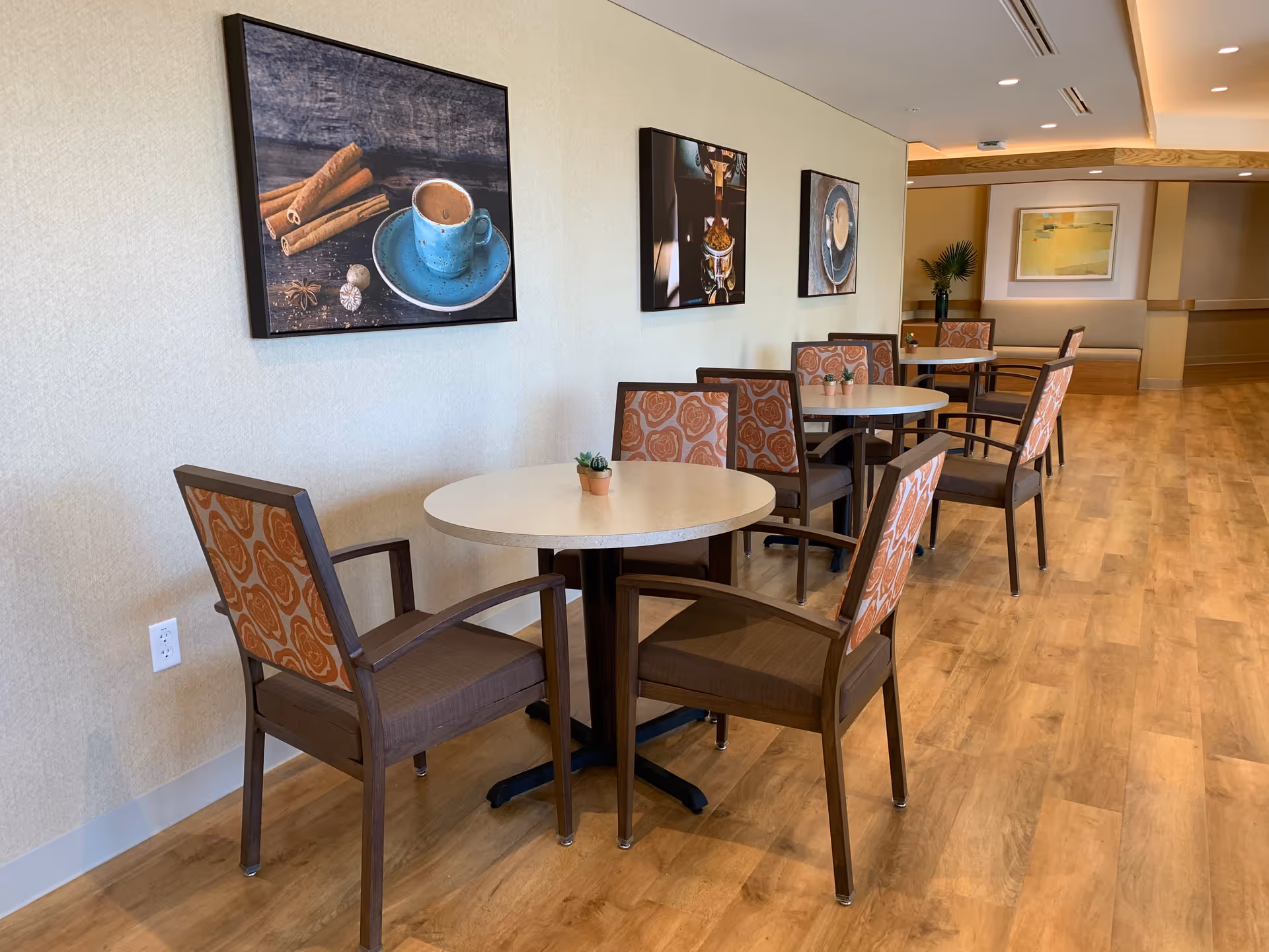 A hallway seating area with round tables and wooden chairs featuring orange patterned cushions. Small potted plants are placed on the tables. The wall has three framed pictures depicting coffee and spices. The floor is wooden, and the hallway extends into a well-lit area with additional seating and artwork on the far wall.