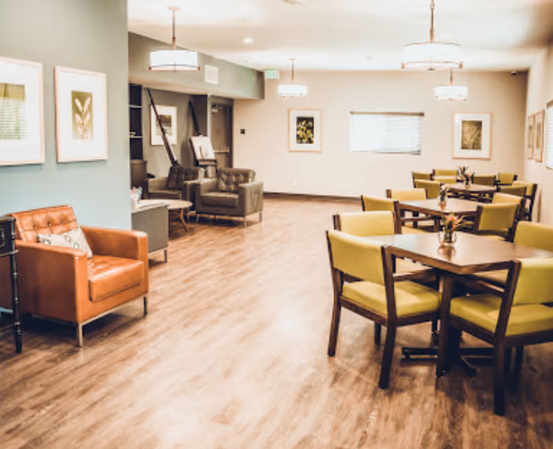 Well-lit communal dining and lounge area with wooden tables and green chairs, leather armchairs, and hardwood floors.