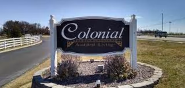 Outdoor view of a black and white sign for Colonial Assisted Living Comm, mounted on white posts surrounded by a circular stone border with small bushes and gravel inside. The sign is located near a paved road with a white fence on the left and open grassy area on the right under a clear blue sky.