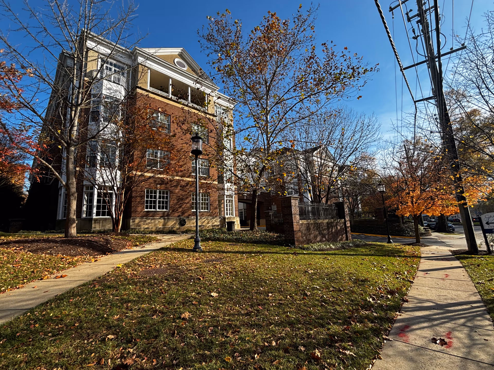 Exterior view of a multi-story brick building with white trim, surrounded by trees with autumn foliage and a grassy lawn. A sidewalk curves through the lawn leading to the building entrance, and a street with parked cars is visible on the right under a clear blue sky.