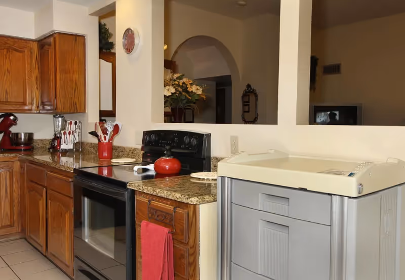 Interior view of a kitchen with wooden cabinets, a black stove with a red kettle on top, granite countertops, a red towel hanging on the oven handle, and various kitchen utensils in holders. There is a clock on the wall and a large gray appliance on the right side. In the background, an archway leads to another room with a floral arrangement and a mirror on the wall.