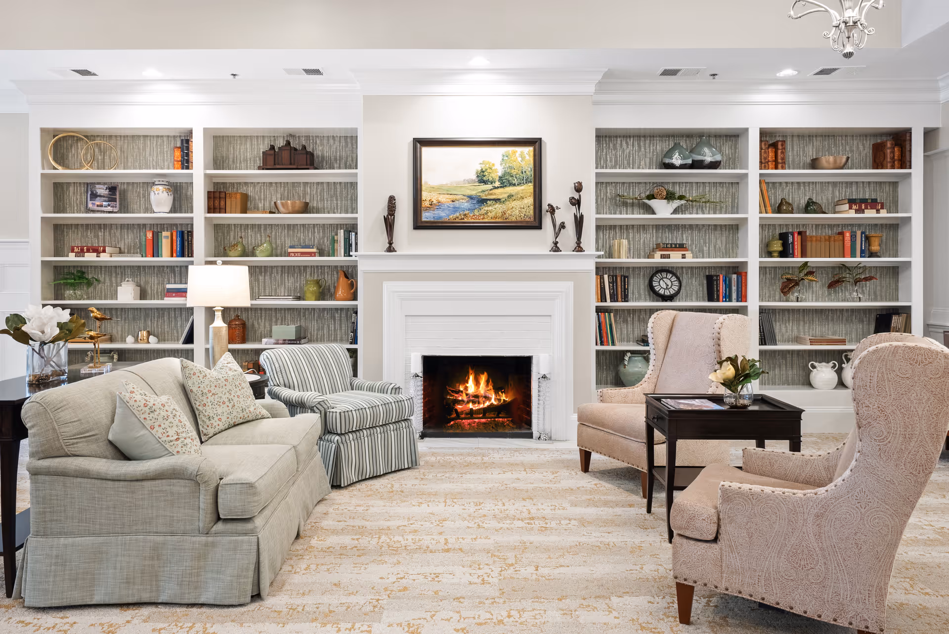 A cozy living room with a lit fireplace in the center, flanked by built-in white bookshelves filled with books and decorative items. The room features a beige sofa with floral pillows, a striped armchair, and two beige wingback chairs around a dark wooden coffee table with a small flower arrangement. The walls are light-colored, and a landscape painting hangs above the fireplace.