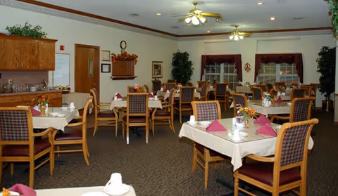 A dining room in a senior living facility with multiple tables covered with white tablecloths, each set with folded napkins, cups, and flower centerpieces. Wooden chairs with cushioned seats and backs surround the tables. The room has ceiling fans with lights, large windows with curtains, and plants placed around the space.