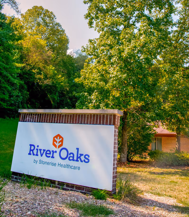 A brick landscape sign reading River Oaks by Stonerise Healthcare in front of trees and a brick building.