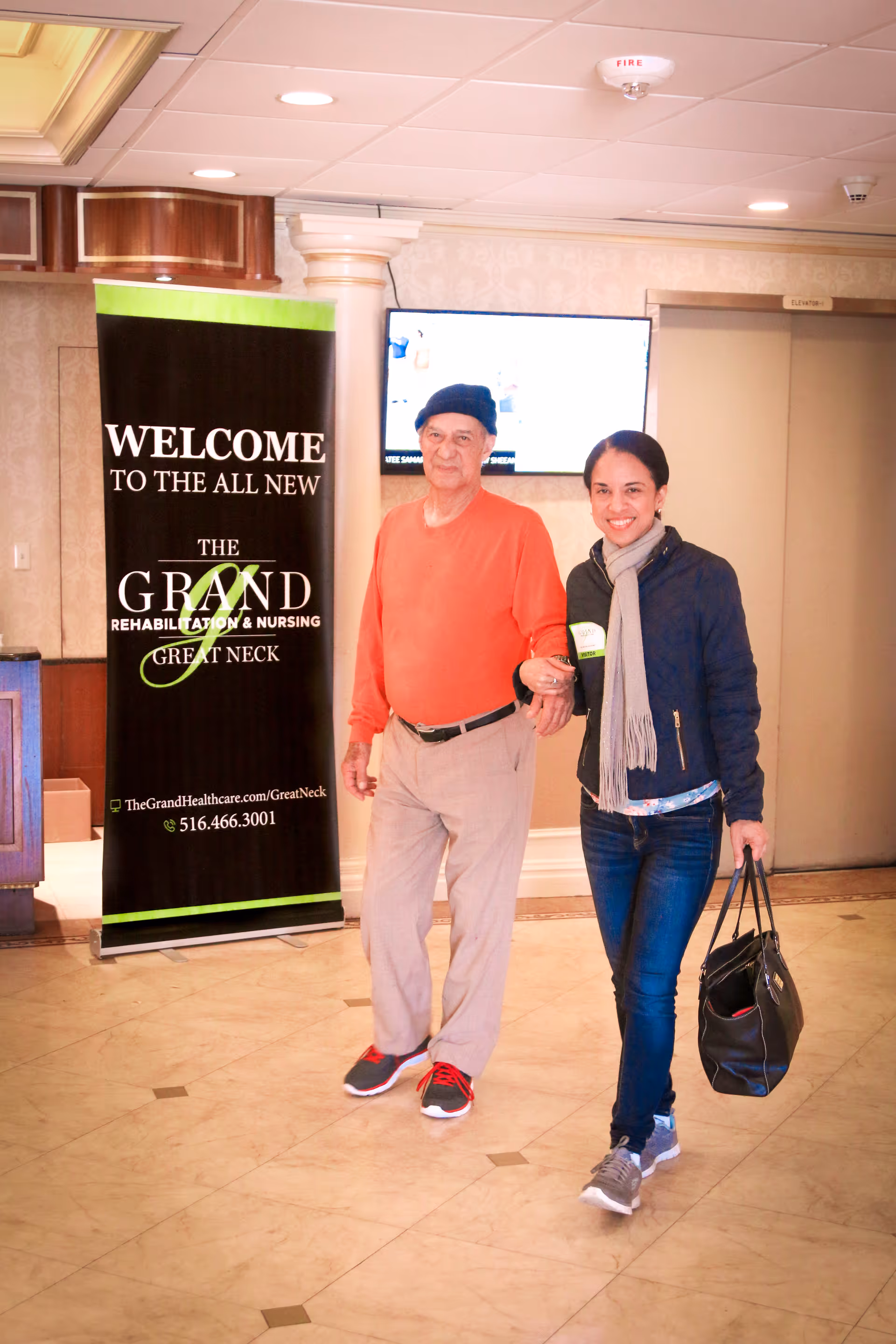An elderly man wearing a black beret, orange sweater, beige pants, and sneakers is walking arm-in-arm with a smiling woman dressed in a dark jacket, scarf, jeans, and sneakers. They are indoors in a well-lit area with a tiled floor. Behind them is a black and green banner that reads 'Welcome to the all new The Grand Rehabilitation & Nursing Great Neck' along with a website and phone number. A TV screen and an elevator door are visible in the background.
