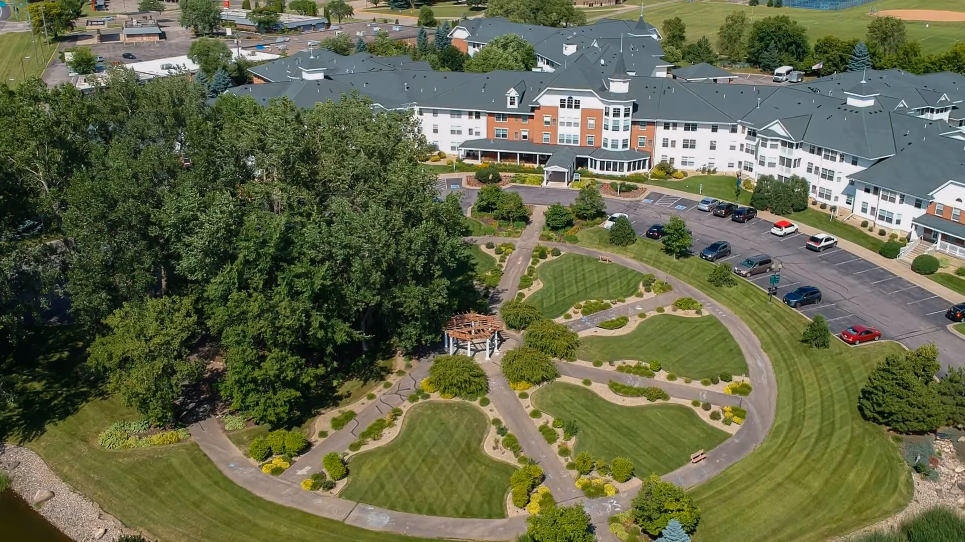 Aerial view of The Rivers Retirement Community showing a large multi-wing building with a green roof surrounded by well-maintained lawns, landscaped gardens, walking paths, and a small gazebo. Several cars are parked in the parking lot adjacent to the building, and there are trees and open green spaces around the property.