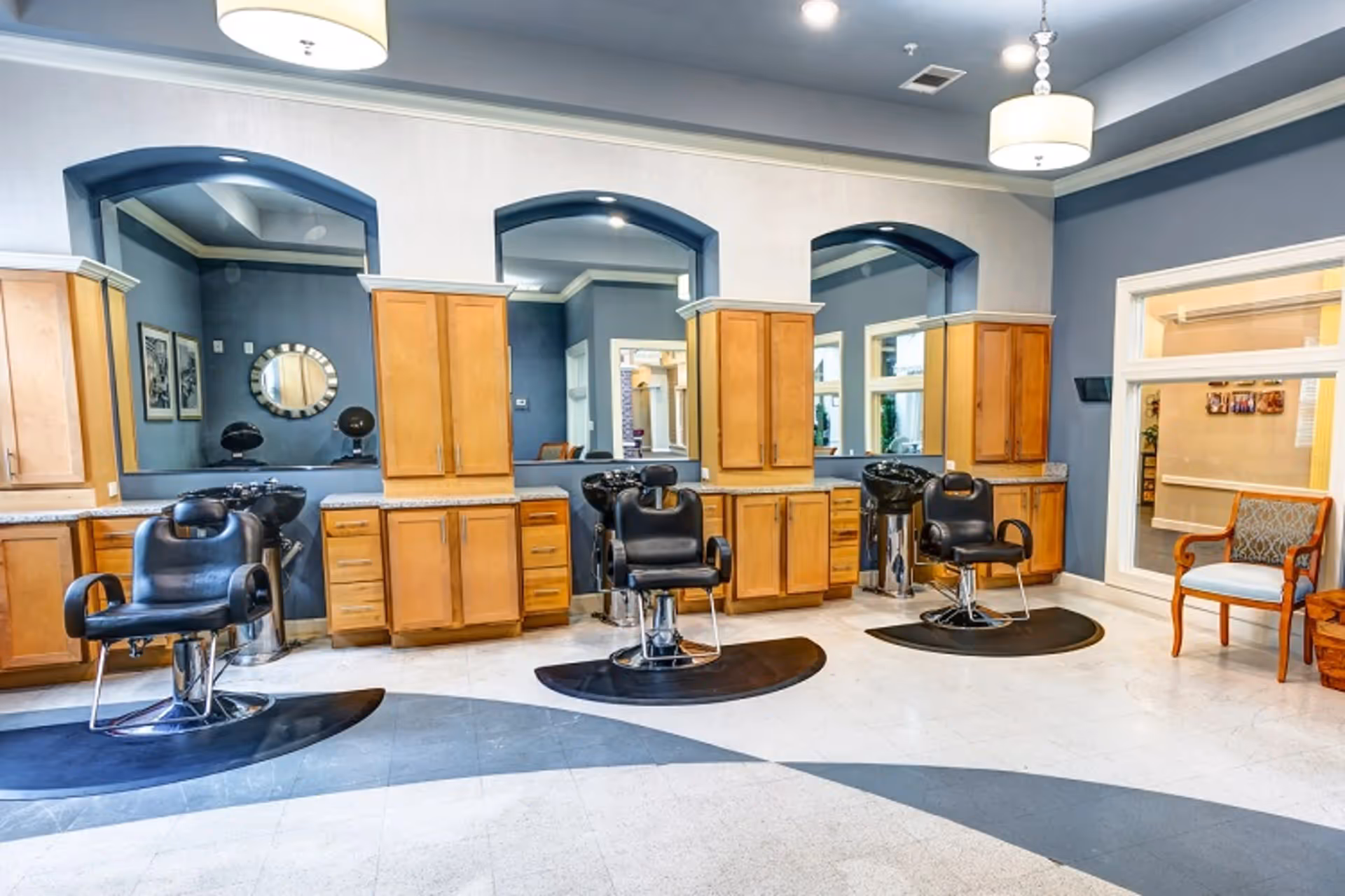 Interior view of a salon area with three black salon chairs in front of large mirrors set into a wall with wooden cabinets. The floor has a curved dark pattern, and there is a single upholstered chair near a window on the right side. The walls are painted blue with white trim and ceiling lights hang from above.